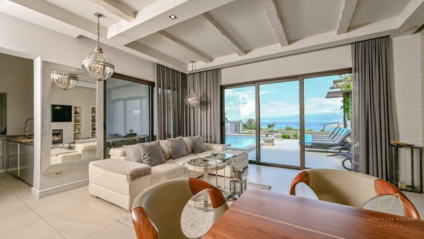 A modern living room with a sofa, dining table, and chairs, a kitchen area, and a large sliding glass door leading to a pool and ocean view. - Seafront Villa Prova 2***** with a top view, indoor & outdoor pool on Krk (Malinska)