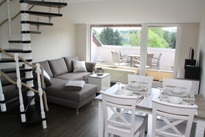 A modern living room with a grey sofa, a dining table set for four, and a spiral staircase. - LOGE 01 | Bright, high-quality 3-room vacation apartment with a stunning view (Braunlage)