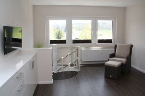 A modern room with a TV, a white cabinet, a wicker chair, and a view of greenery through large windows. - LOGE 01 | Bright, high-quality 3-room vacation apartment with a stunning view (Braunlage)