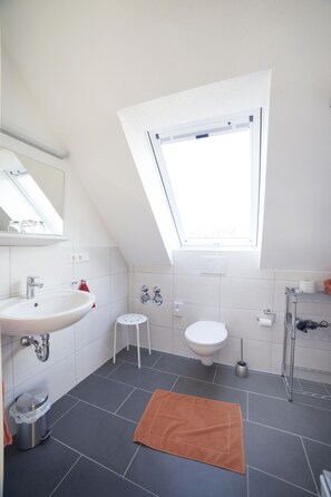 Un baño moderno con tragaluz, azulejos blancos, lavamanos, inodoro y un taburete pequeño.