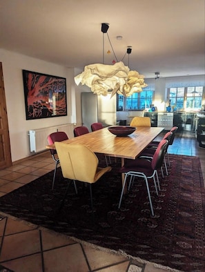 Een eetruimte met een houten tafel, rode en gele stoelen, een groot vloerkleed en een bijzondere hanglamp.
