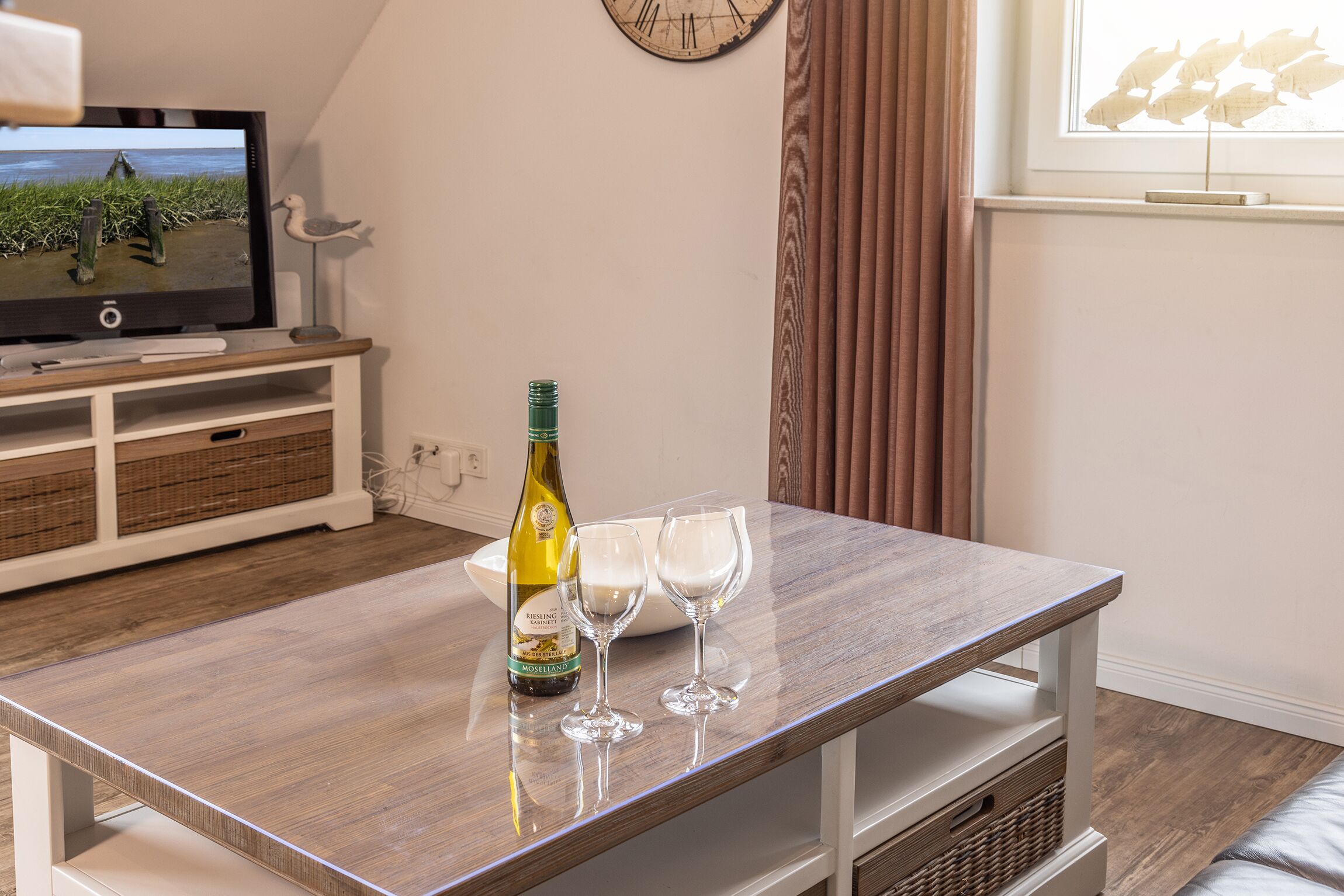 A wooden table with a bottle of wine and two glasses, a television, wicker baskets, and a clock on the wall.