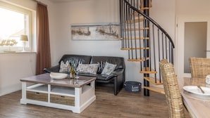 Een moderne woonkamer met een zwarte leren bank, een witte salontafel, een schilderij aan de muur, een trap met een zwarte leuning en rieten stoelen bij een eettafel.