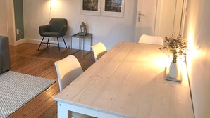 Ein Essbereich mit einem Holztisch, weißen Stühlen, einem kleinen Beistelltisch mit einer Laterne und zwei gerahmten Bildern an der Wand.