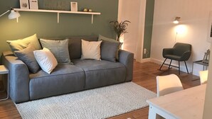 Ein Wohnzimmer mit grauem Sofa, weißen Kissen, einem weißen Teppich, einem kleinen Tisch und einer Pendelleuchte.