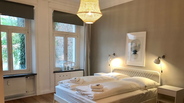 Ein Schlafzimmer mit Bett, Nachttischen, einer Hängelampe, einem Fenster mit Blick auf Grünflächen und einer Heizung.