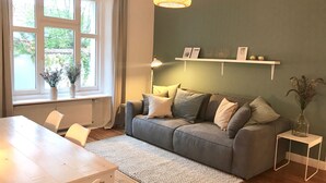 Ein Wohnzimmer mit grauem Sofa, weißen Kissen, einem kleinen weißen Tisch und einem Fenster mit Vorhängen.