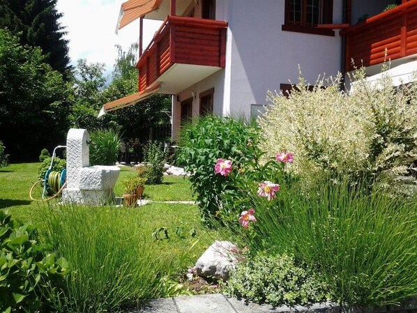 A house with a balcony, a garden with flowers and plants, and a stone structure.