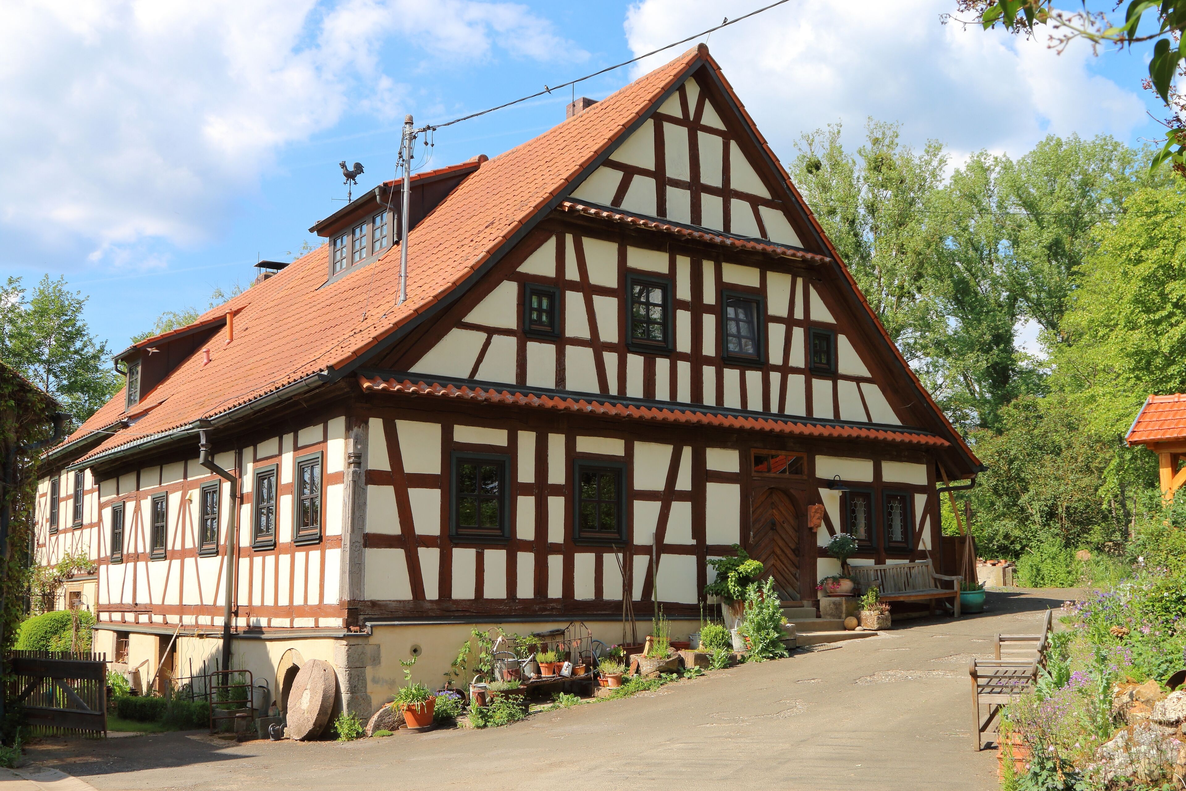 Ein traditionelles Fachwerkhäuschen mit geneigtem Dach, einer Holztür und einem kleinen Vorbereich mit Blumentöpfen.