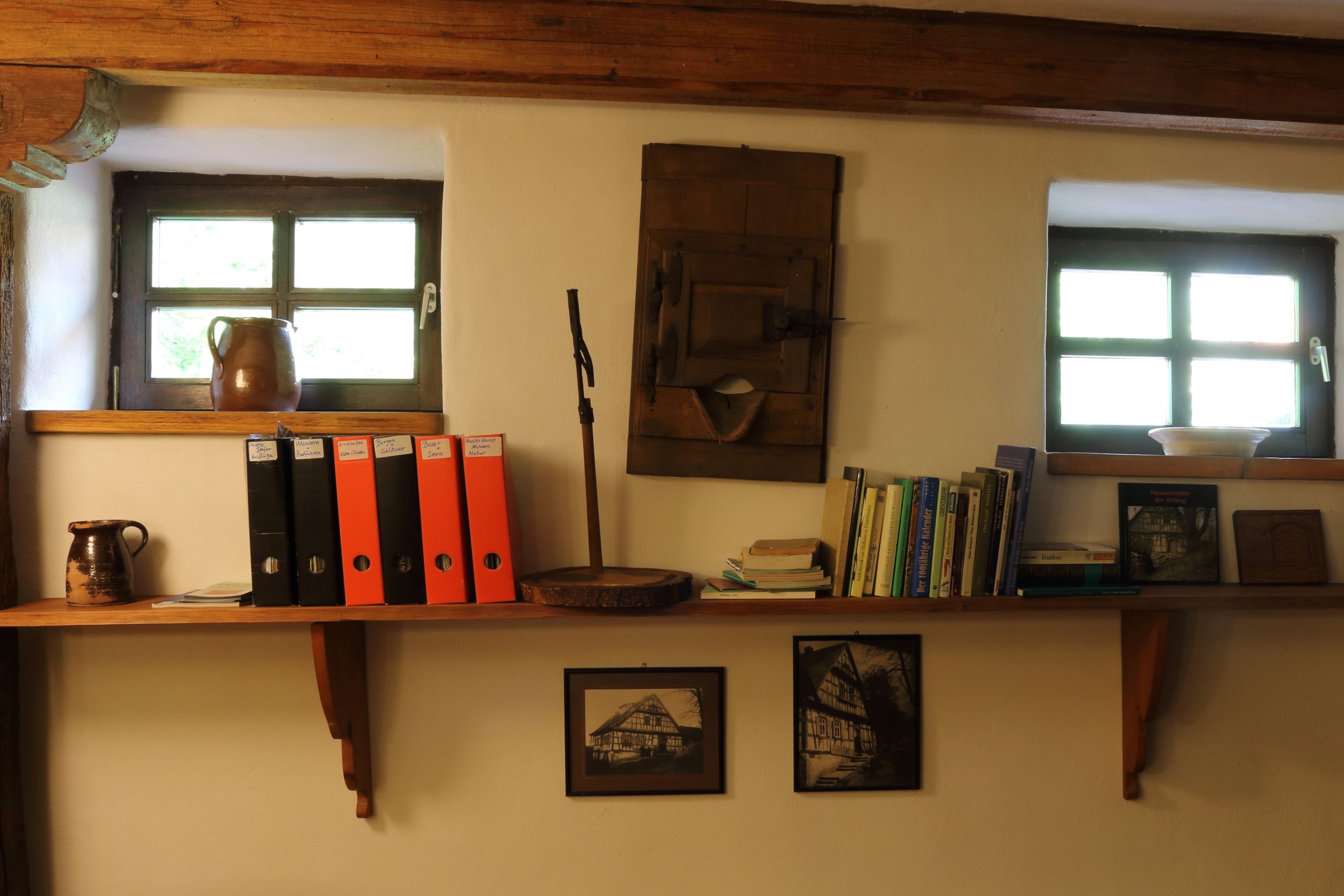 A room with a wooden shelf holding books, binders, and a ceramic pitcher. There are two windows with curtains, one with a view of greenery outside.