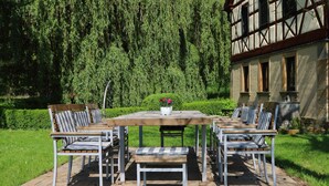 An outdoor patio with a wooden table and six chairs, surrounded by a well-maintained garden and a traditional building in the background. - Holiday in 300 years old, listed mill in an idyllic location (Hofheim in Unterfranken)