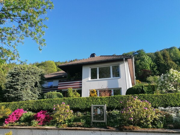 A two-story house with a sloped roof, surrounded by lush greenery and flowering plants.