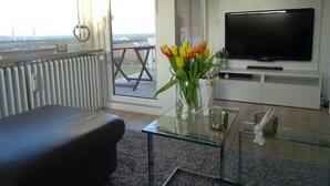 Ein modernes Wohnzimmer mit einem Glastisch, einer schwarzen Ledersofa, einem Flachbildfernseher auf einer weißen Unterhaltungsanlage und Meerblick durch große Fenster.