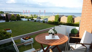 Ein Balkon mit einem runden Holztisch, weißen Stühlen und einem kleinen Tisch in Weiß. Im Hintergrund ist Meerblick und eine Flaggenwehe zu sehen.