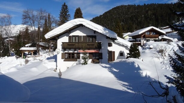 Un chalet enneigé, doté d’une façade en bois, entouré d’arbres et de montagnes également recouverts de neige.