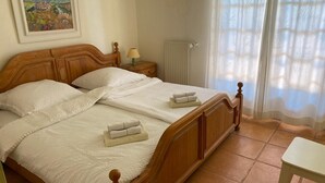 A double bed with a wooden frame, two pillows, and two folded towels on the bed. There is a nightstand with a lamp, a radiator, and a window with sheer curtains. - Holiday villa with pool, large terrace and great sea and far view. (Gattières)