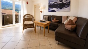 A living room with a sofa, a wicker chair, a table, and a painting of a mountain landscape. - Cozy, sunny & newly renovated apartment with a view of the Zugspitze - 4 stars (Garmisch - Partenkirchen)