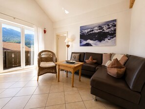 A living room with a sofa, a wicker chair, a table, and a painting of a mountain landscape.