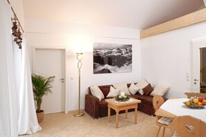 A cozy living room with a brown sofa, a wooden coffee table, and a dining table with chairs. A potted plant is near the entrance, and a floor lamp is beside the sofa.
