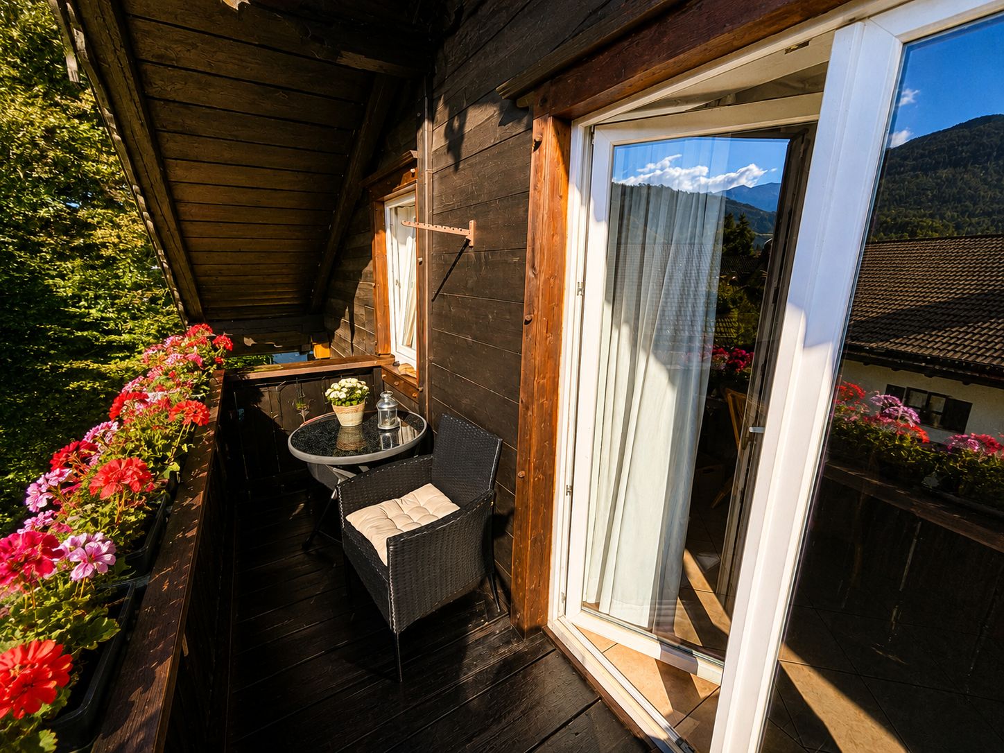 Eine Holzdiele mit Glastür und Blick auf die Berge, ein kleiner Tisch mit einem Stuhl sowie Blumenkästen an der Wand.