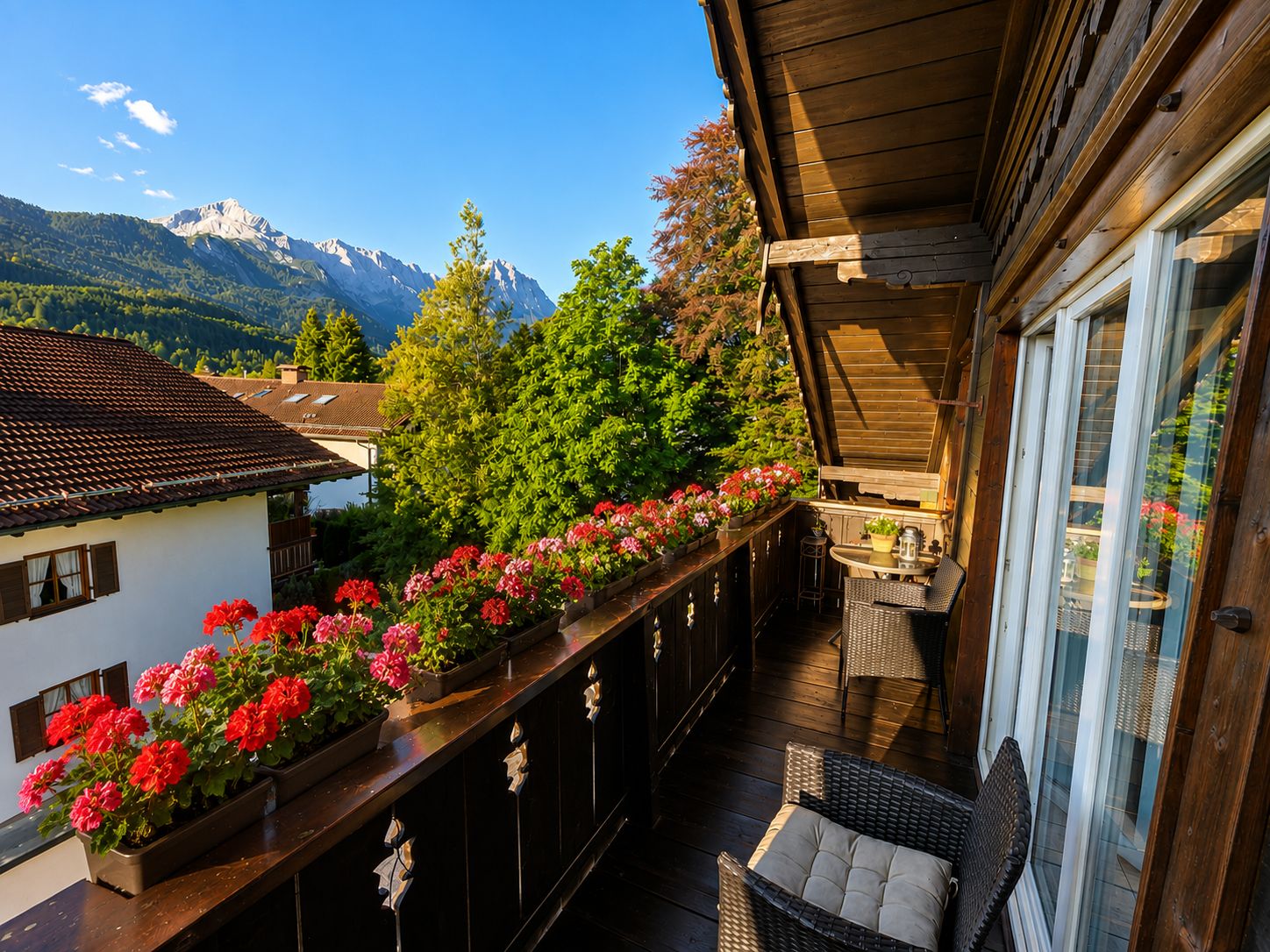 A balcony with a wooden railing, potted flowers, wicker furniture, and a view of mountains.