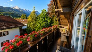 A balcony with a wooden railing, potted flowers, wicker furniture, and a view of mountains.