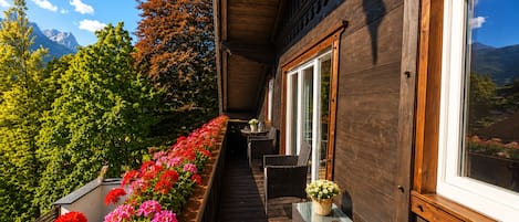A wooden balcony with a row of red flowers, wicker furniture, and a view of mountains.