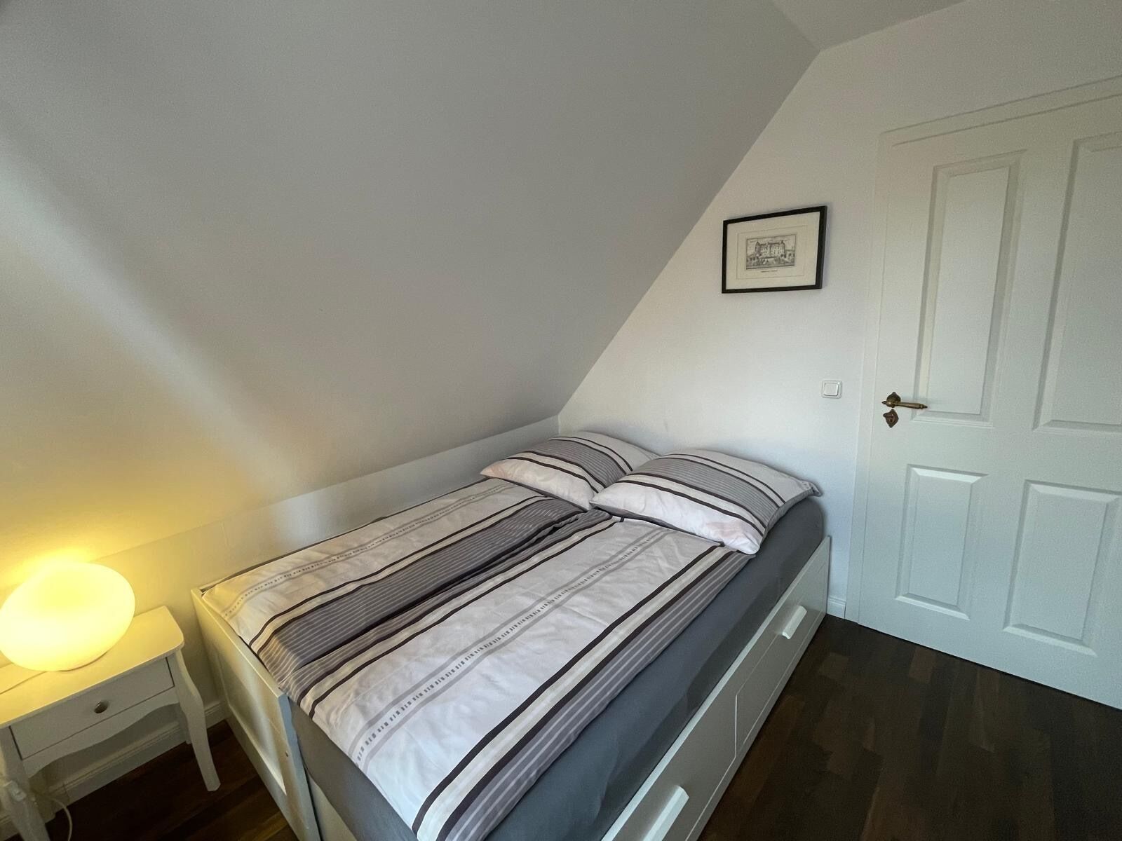Ein kleines, ordentlich bezogenes Bett mit Streifenmuster in einem Zimmer mit weißen Wänden und einer Tür.