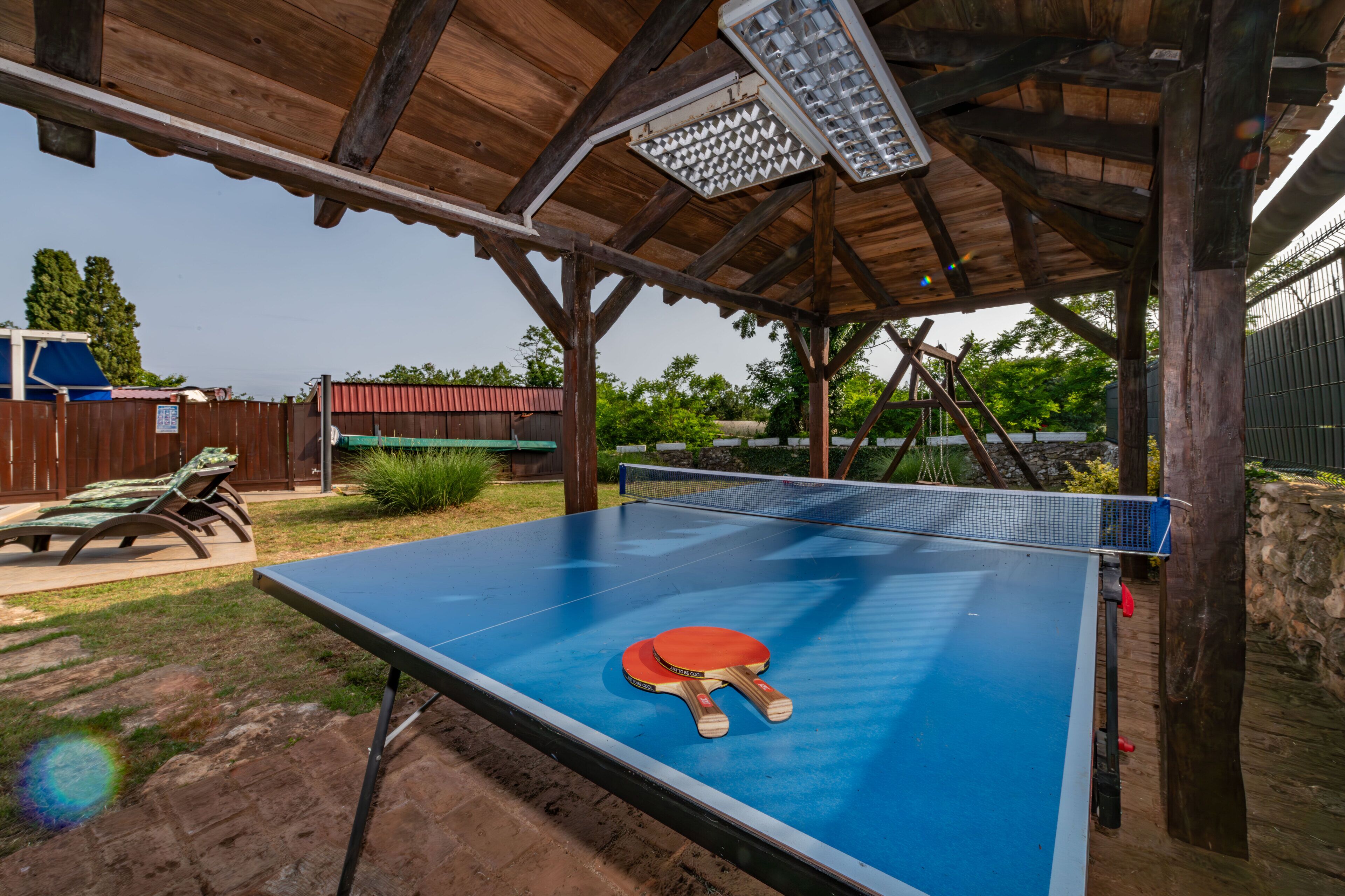 An outdoor ping pong table with a net, surrounded by wooden beams and a metal roof structure.