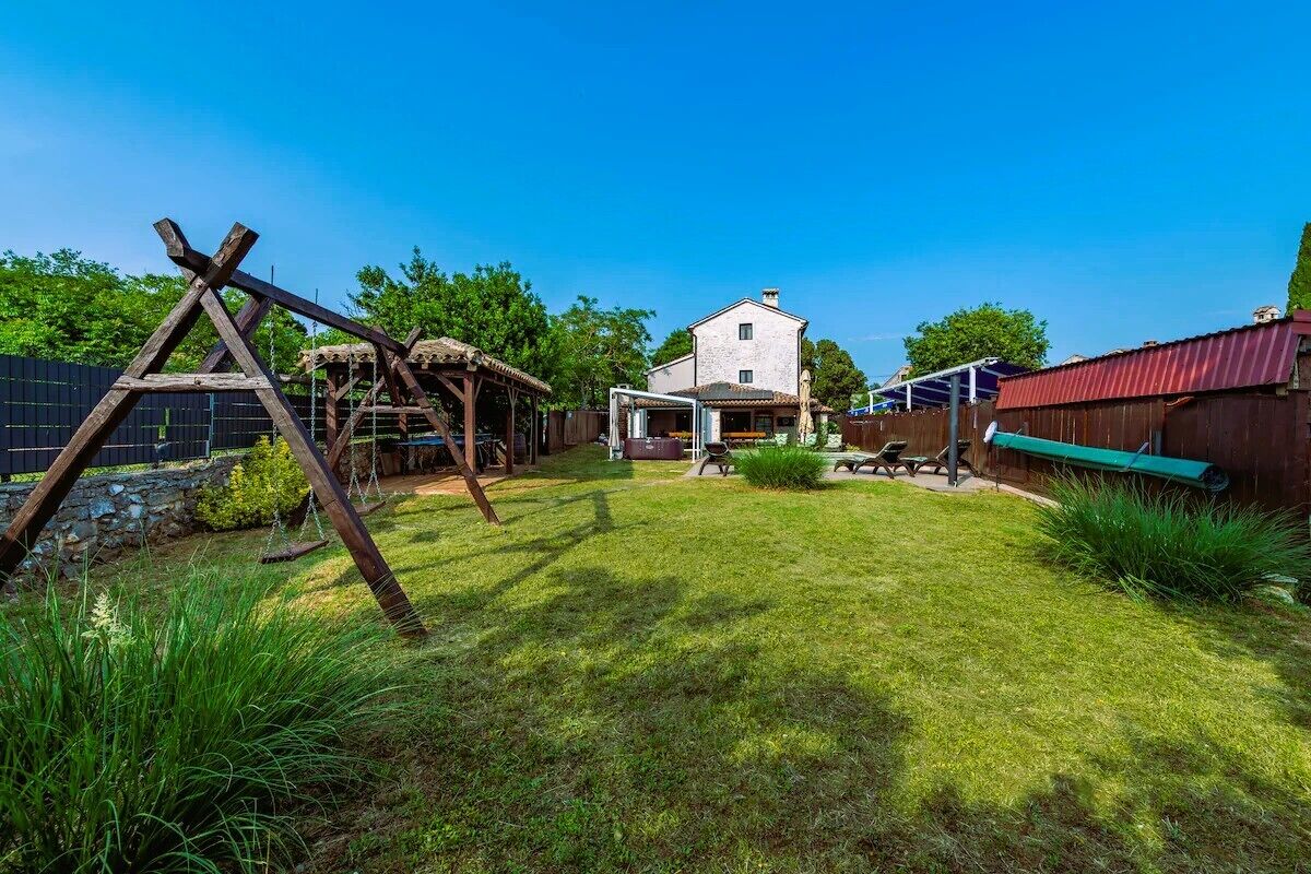 A backyard with a wooden swing set, a covered patio area, and a stone wall.