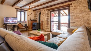 Ein gemütliches Wohnzimmer mit Steinwand, Holzbalken, Fernseher, Kamin und bequemen Sitzmöglichkeiten.