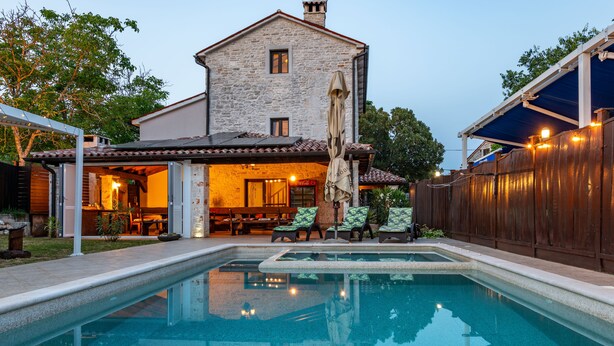 Ein Schwimmbecken mit einem dahinterliegenden Steinhaus, einer überdachten Terrasse und Sitzgelegenheiten im Freien.