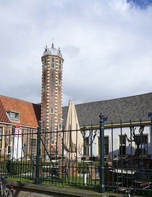 Une tour historique dotée d’une horloge, entourée d’un espace extérieur aménagé avec des chaises et des tables, le tout délimité par une clôture.