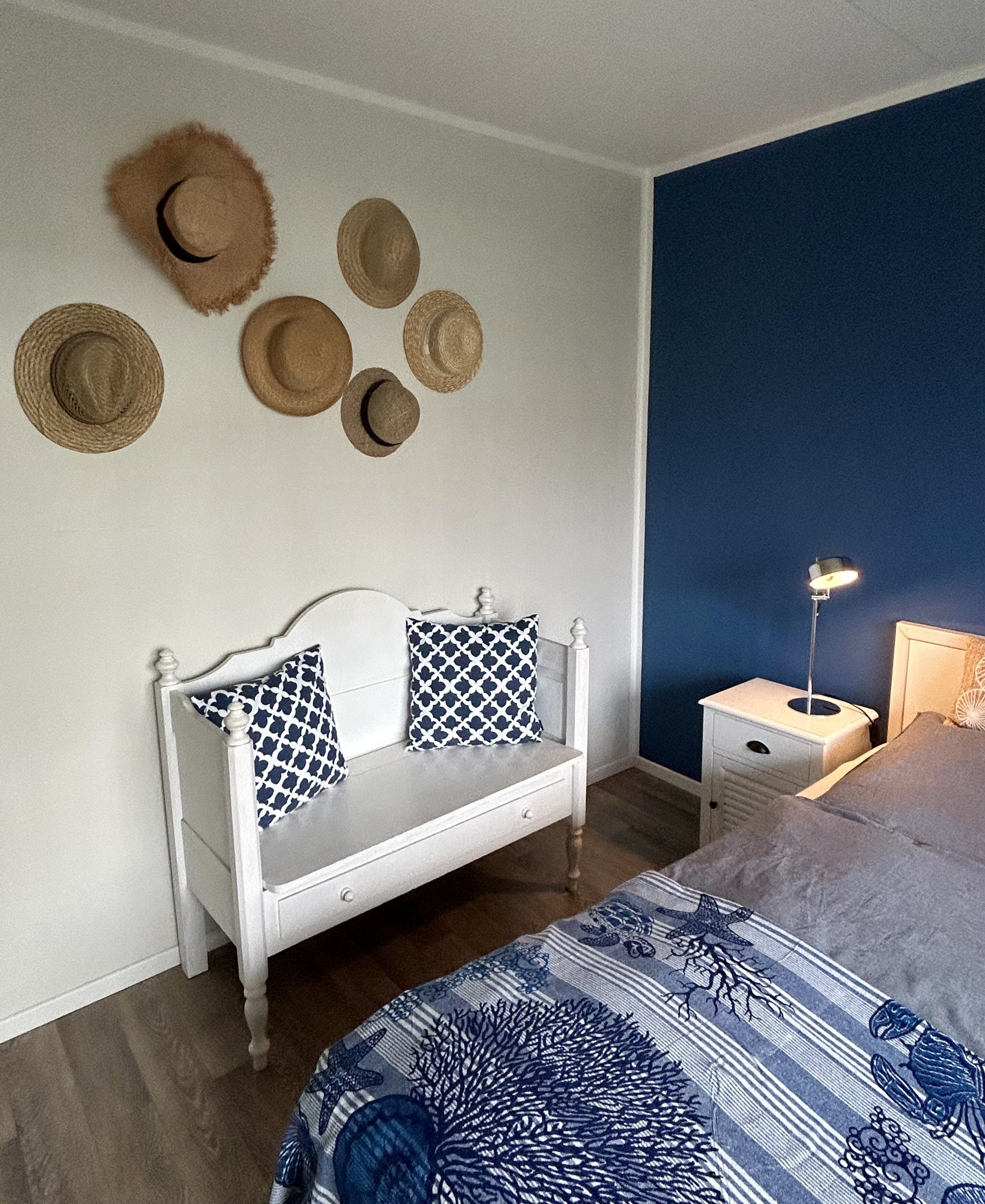 Un dormitorio con una cama blanca, un banco blanco con almohadas estampadas, una mesita de noche con lámpara y varios sombreros en la pared.