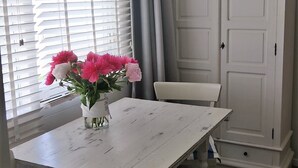 A white dining table with a vase of pink flowers, a striped cushion, and a white cabinet in a room with wooden flooring.