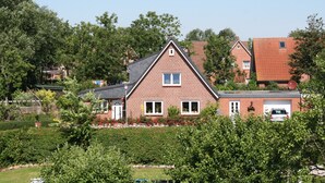 Une maison à deux étages, construite en briques et dotée d’un toit à pignon, entourée d’une végétation luxuriante et d’arbres.