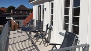 A covered outdoor area with a table and chairs, a wooden deck, and a white building with large windows. - Bright & modern, 4-star apartment for up to 5-6 people (maisonette) (Langeoog)