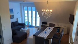 A modern living room with a dark sofa, a dining table with chairs, a chandelier, and a large window with blinds. - Bright & modern, 4-star apartment for up to 5-6 people (maisonette) (Langeoog)