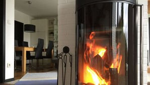 A modern living room with a glass-fronted fireplace, a dining area with a wooden table and chairs, and a white brick wall. - Beach and sea(h)r in a vacation home with garden close to the beach at Schilksee⛵️ (KI)