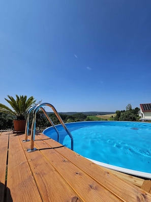An outdoor swimming pool with a wooden deck and metal ladder, surrounded by greenery and a clear sky.
