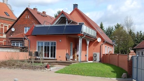 Ein Wohnhaus mit roter Backsteinfassade, einem Balkon mit Solaranlage, einer Haustür und einem kleinen Vorgarten mit Holzzaun.
