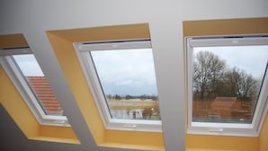 Ein Zimmer mit drei Oberlichtern, jeweils mit weißem Rahmen und gelbem Sockel, mit Blick auf eine Wohngegend mit Häusern und Bäumen.