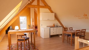 Ein gemütlicher, gut beleuchteter Dachbodenraum mit einem Holzesstisch, Stühlen, einem kleinen Kühlschrank und einer Küchenzeile mit Schränken und Spüle.