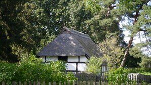 Une maison traditionnelle à toit de chaume, entourée d’une clôture en bois et d’une végétation luxuriante.