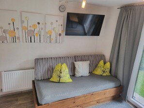 A cozy living room with a grey sofa, yellow and white cushions, a flat-screen TV mounted on the wall, and a window with grey curtains.