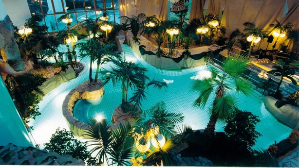 Un área de piscina interior con múltiples fuentes, palmeras e iluminación ambiental.