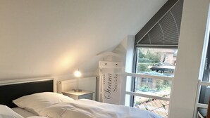 A bedroom with two beds, a nightstand, a lamp, a radiator, and a view of the outside. - Studio apartment in restored captain's house with a panoramic view of the port (Krummhörn)