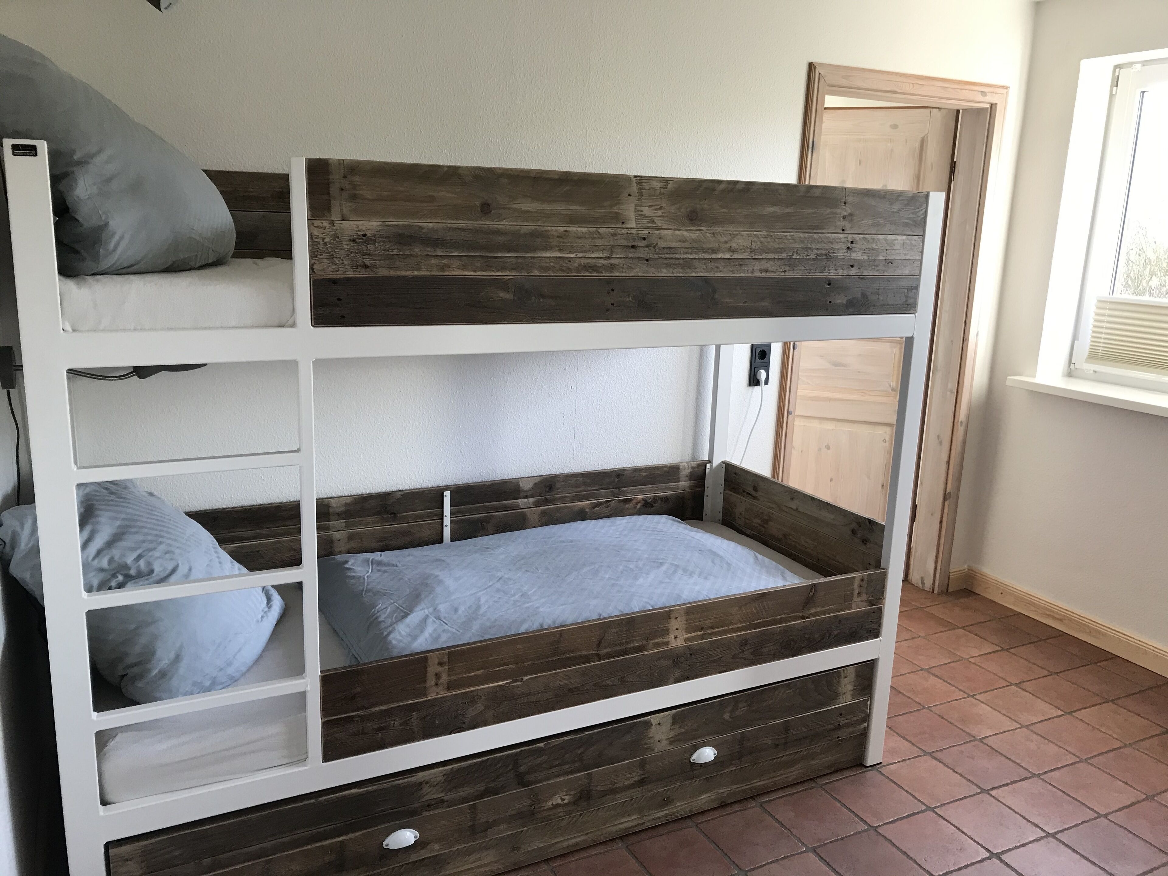 Een stapelbed met drie niveaus, elk met een kussen en een matras. Er is een houten deur en een raam met zonwering.