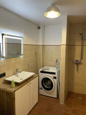 A compact bathroom with a sink, mirror, washing machine, and shower.