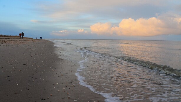 A beach with a wide sandy shore, gentle waves, and a few people walking along the water's edge.